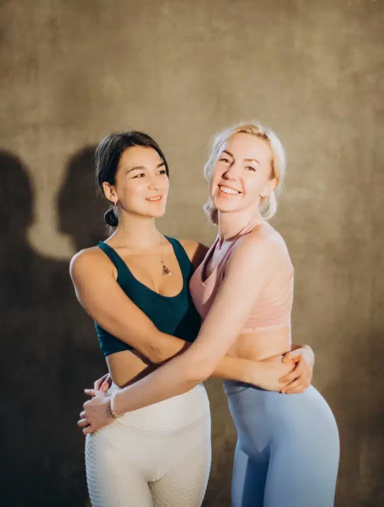 Two women smiling and hugging each other in casual workout attire against a warm-toned background.