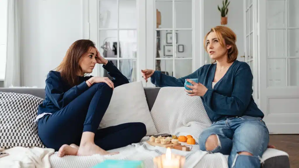 Two women sitting on a sofa at home, having a relaxed conversation while drinking and sharing snacks on a table.