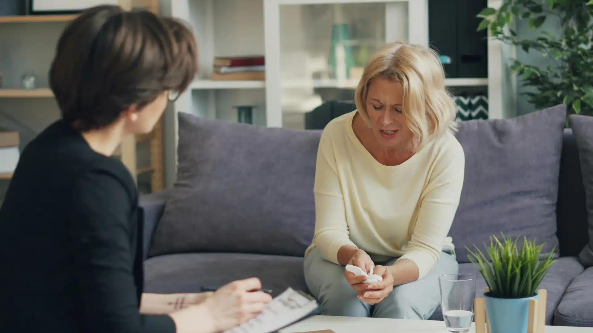 A woman sitting on a couch talking to a therapist, holding a tissue while the therapist takes notes during a counseling session.