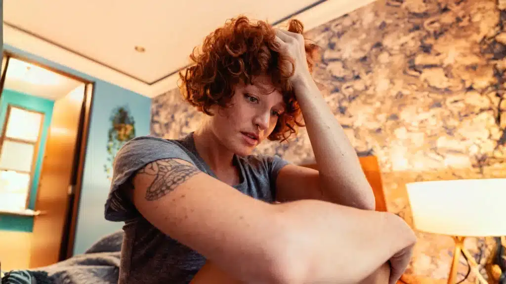 Woman sitting on bed with hand in hair, looking anxious and deep in thought about managing anxiety.