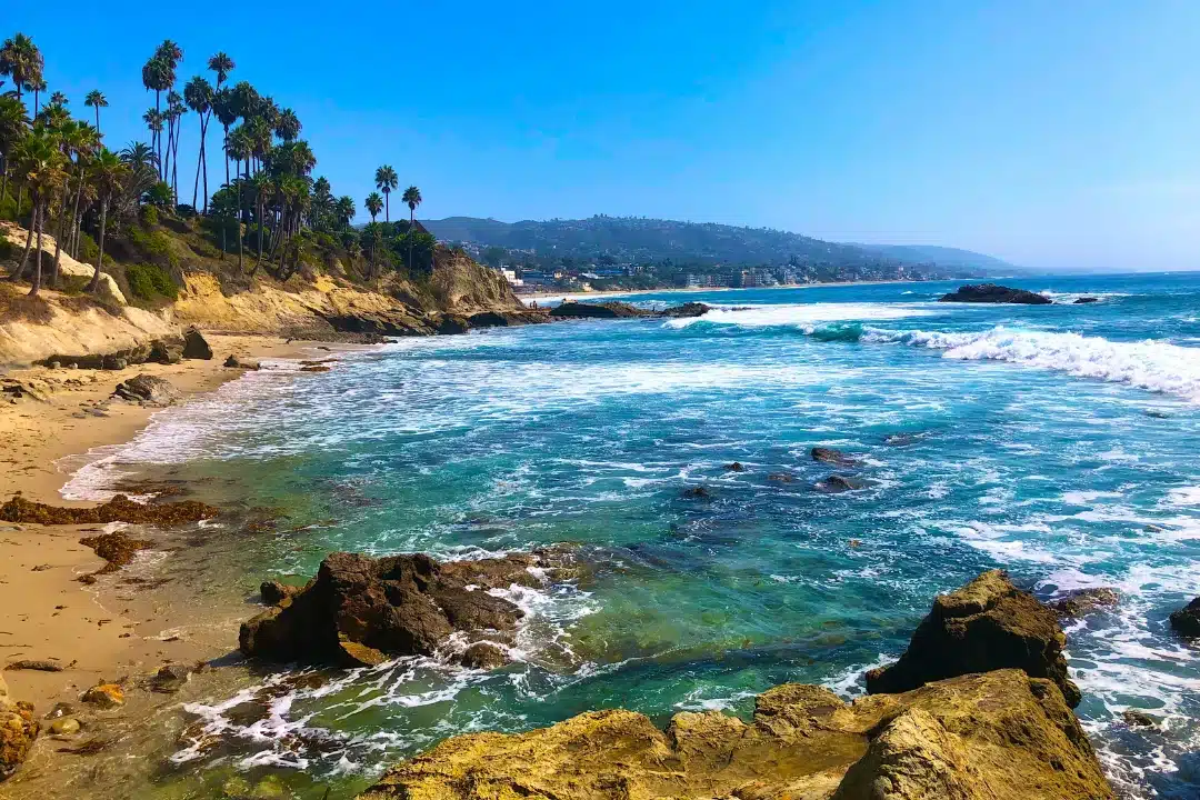 Rocky shoreline and clear blue waves along the coast of Laguna Beach with cliffs and palm trees overlooking the ocean.