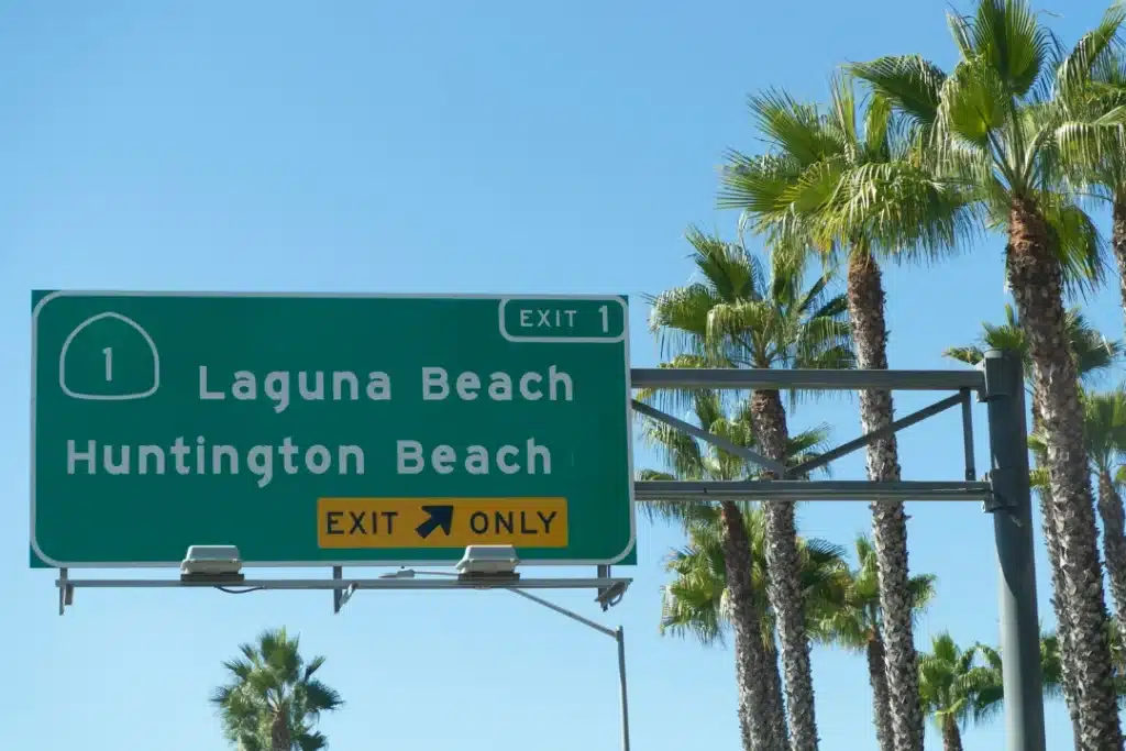Laguna Woods road sign near palm trees pointing toward nearby beach destinations under bright daylight